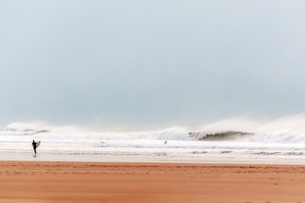 Strong offshore wind with large waves at Croyde Beach in Croyde Bay. Surfer with surf board in wetsuit walking along the sand.
